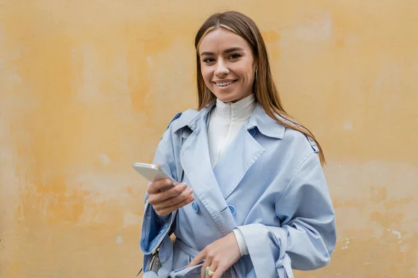 Cheerful woman in blue trench coat using smartphone near weathered yellow wall on street in Vienna — Stock Photo