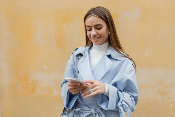 Happy woman in blue trench coat messaging on smartphone near weathered yellow wall on street in Vienna — Stock Photo