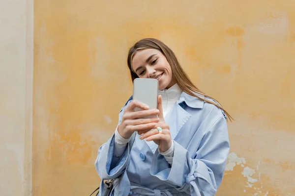 Cheerful woman in blue trench coat taking photo on smartphone near weathered yellow wall on street in Vienna — Stock Photo