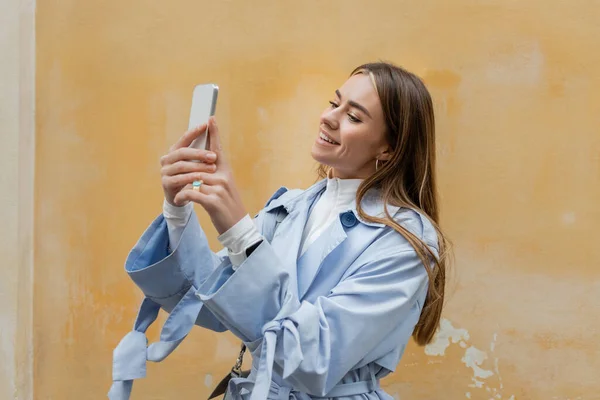 Happy woman in blue trench coat taking photo on smartphone near weathered yellow wall on street in Vienna — Stock Photo