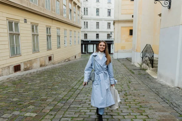 Happy young woman in blue trench coat walking with shopping bags on street in Vienna — Stock Photo