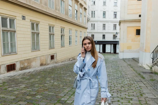 Mujer joven con gabardina azul hablando por teléfono inteligente mientras camina por la calle en Viena - foto de stock
