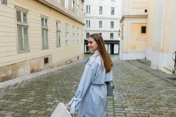 Happy young woman in blue trench coat walking near buildings on street in Vienna — Stock Photo