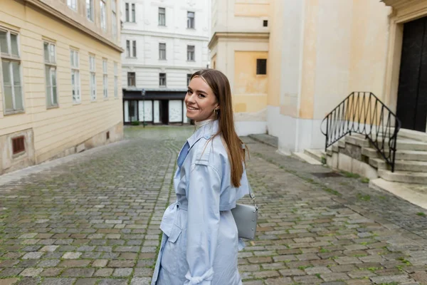Cheerful young woman in blue trench coat walking near buildings on street in Vienna — Stock Photo
