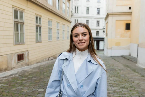 Cheerful young woman in blue trench coat standing near buildings on street in Vienna — Stock Photo