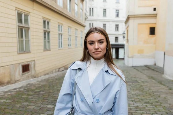 Charming young woman in blue trench coat standing near buildings on street in Vienna — Stock Photo