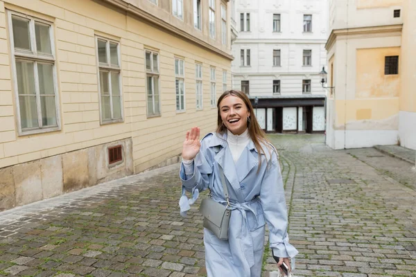 Charming woman in blue trench coat waving hand while walking with smartphone near buildings in Vienna — Stock Photo