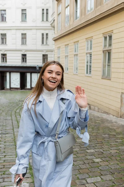 Happy woman in blue trench coat waving hand while walking with smartphone near buildings in Vienna — Stock Photo