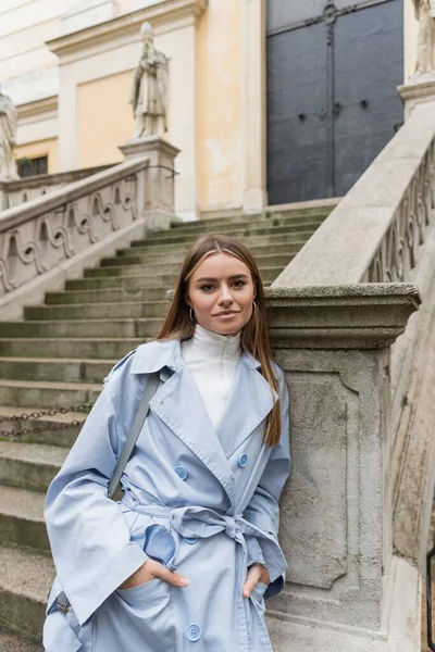 Encantadora mulher em casaco de trincheira azul de pé com as mãos em bolsos perto de escadas antigas na rua em Viena — Fotografia de Stock