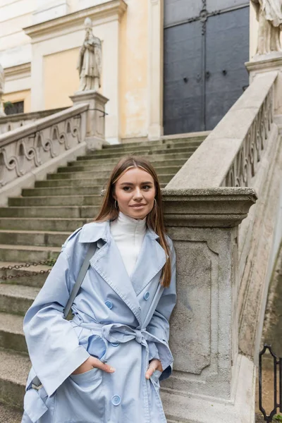 Charming woman in blue trench coat smiling while standing with hands in pockets near ancient stairs on street in in Vienna — Stock Photo