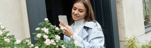 Happy young woman in blue trench coat taking photo of green bush with blooming flowers on street in Vienna, banner — Stock Photo