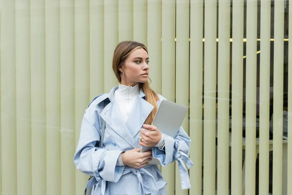 Pretty woman in blue trench coat holding laptop while standing near window on street in Vienna — Stock Photo