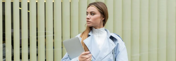 Pretty woman in blue trench coat holding laptop while standing near window shades on street in Vienna, banner — Stock Photo