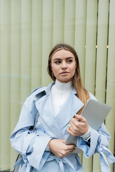 Pretty freelancer in blue trench coat holding laptop while standing near window on street in Vienna — Stock Photo