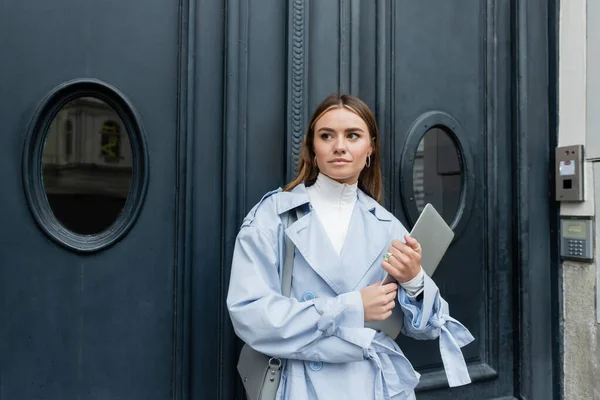 Pretty freelancer in blue trench coat holding laptop while standing on street in Vienna — Stock Photo