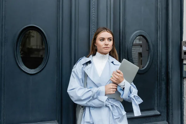 Young freelancer in blue trench coat holding laptop while standing on street in Vienna — Stock Photo
