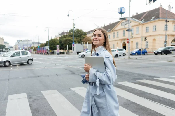 Glückliche Frau im blauen Trenchcoat hält Laptop in der Hand und überquert Straße in Wien — Stockfoto