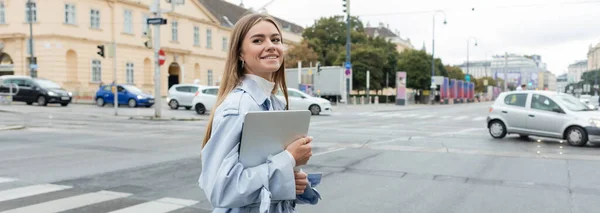 Happy woman in blue trench coat holding laptop and crossing road on street in Vienna, banner — Stock Photo