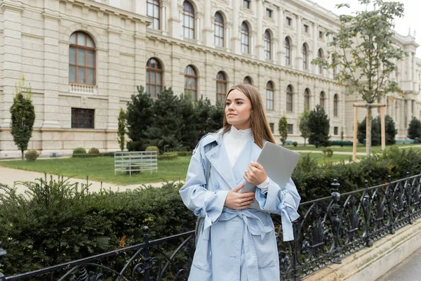 Mujer joven con gabardina azul sosteniendo portátil cerca del edificio histórico en Viena - foto de stock