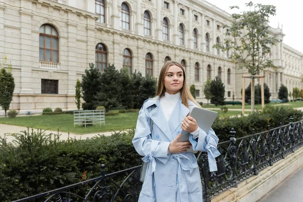 Encantadora jovem mulher em casaco de trincheira azul segurando laptop perto de edifício histórico em Viena — Fotografia de Stock