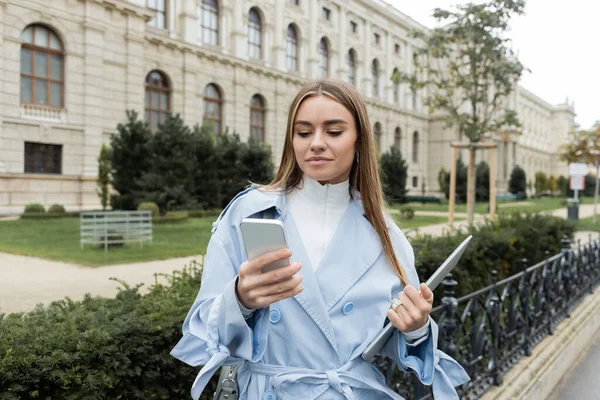 Молода жінка в синьому плащі з ноутбуком і смартфоном біля історичного будинку у Відні. — стокове фото