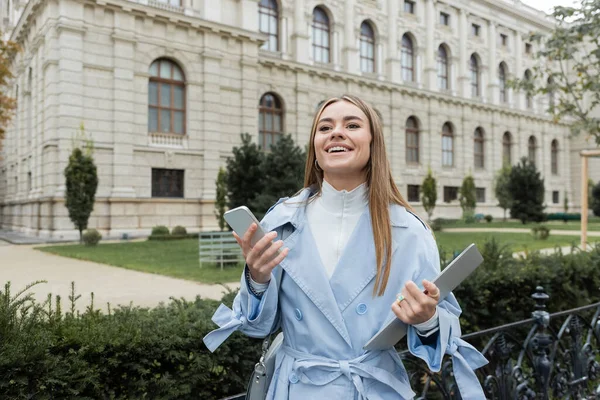 Счастливая молодая женщина в голубом плаще с ноутбуком и смартфоном возле исторического здания в Вене — стоковое фото