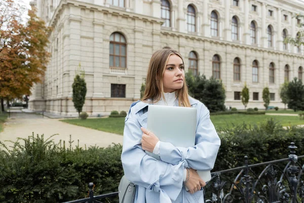 Bonita jovem mulher em casaco de trincheira azul segurando laptop perto de edifício histórico em Viena — Fotografia de Stock