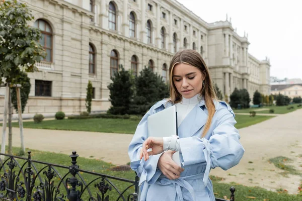 Pretty woman in blue trench coat holding laptop and checking time on wristwatch near historical building in Vienna — Stock Photo