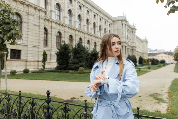 Pretty woman in blue trench coat checking time on smart watch while waiting near historical building in Vienna — Stock Photo
