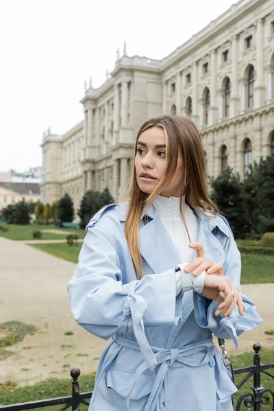 Mujer joven con gabardina azul comprobando el tiempo en reloj de pulsera mientras espera cerca del edificio histórico de Viena - foto de stock