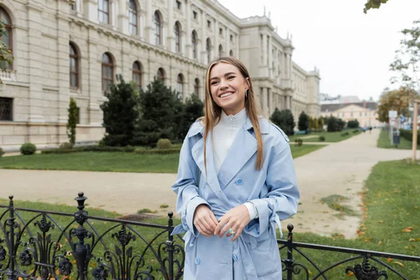 Joyful young woman in blue trench coat standing near historical building in Vienna — Stock Photo