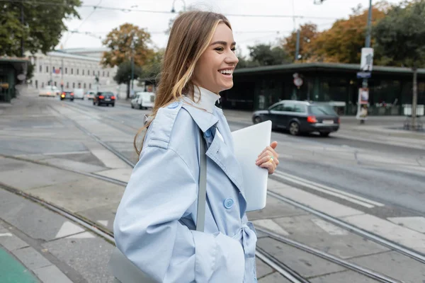 Fröhliche Frau im blauen Trenchcoat mit Laptop beim Überqueren der Straße in Wien — Stockfoto