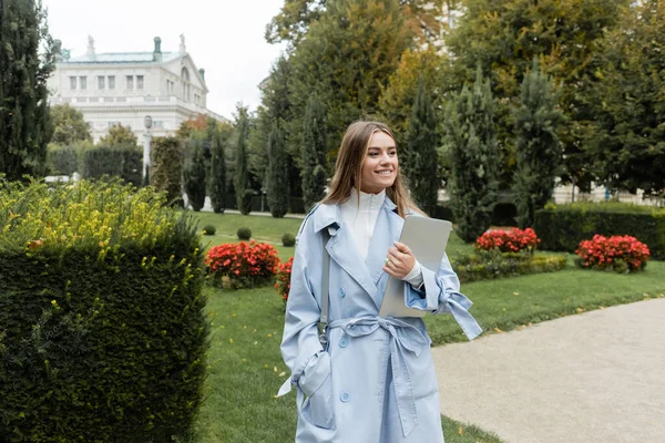 Cheerful woman in blue trench coat holding laptop while standing with hand in pocket in green park in Vienna — Stock Photo