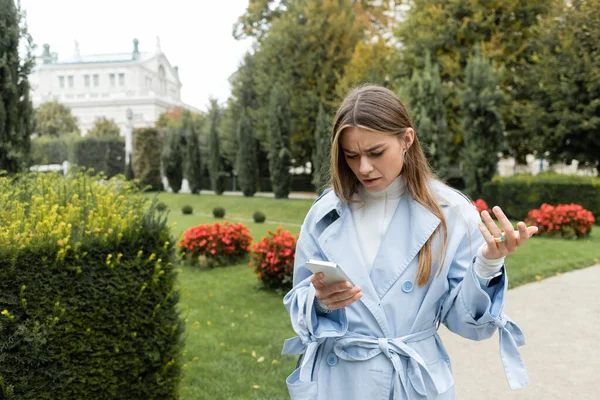 Worried young woman in blue trench coat using smartphone while standing in green park in Vienna — Stock Photo