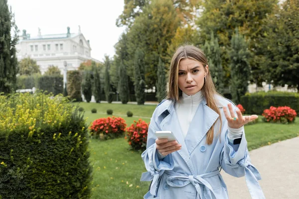 Confused woman in blue trench coat using smartphone while standing in green park in Vienna — Stock Photo