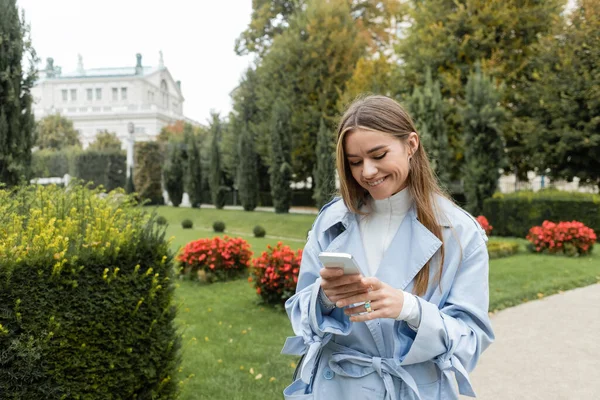 Smiling young woman in blue trench coat using smartphone while standing in green park in Vienna — Stock Photo