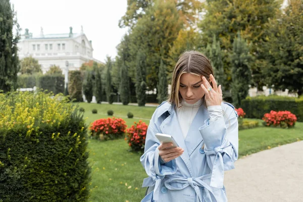 Giovane donna in trench blu con cellulare in piedi nel parco verde di Vienna — Foto stock
