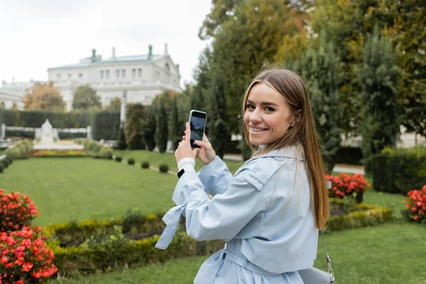 Happy young woman in blue trench coat taking photo of green park on smartphone — Stock Photo