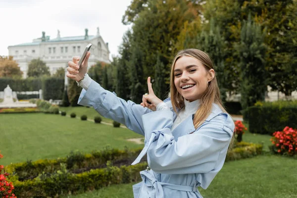 Mujer joven y feliz con gabardina azul apuntando hacia el exterior mientras usa el teléfono inteligente en el parque verde de Viena - foto de stock