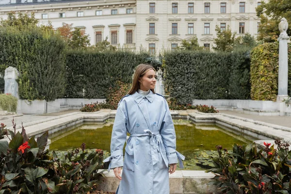 Молодая женщина в синем плаще, стоящая возле фонтана и исторического здания в зеленом парке в Вене — стоковое фото