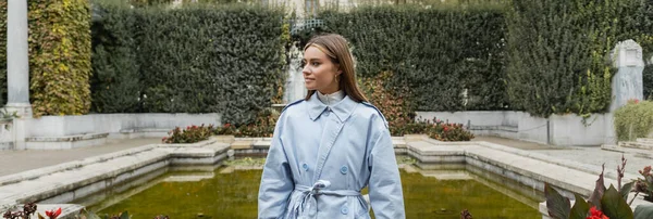 Young woman in blue trench coat standing near fountain with green water in park, banner — Stock Photo