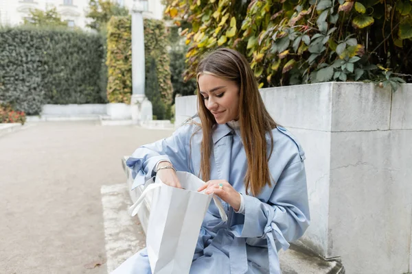 Fröhliche Frau im blauen Trenchcoat auf Betonbank sitzend und in Papiertüte im grünen Park in Wien schauend — Stockfoto