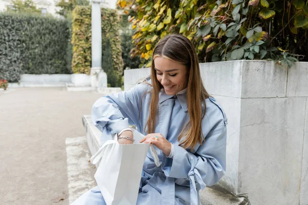 Zufriedene Frau im blauen Trenchcoat sitzt auf Betonbank und blickt in grüne Einkaufstüte im Park in Wien — Stockfoto