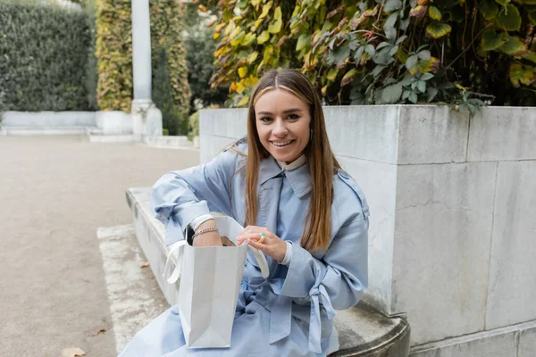 Glückliche junge Frau im blauen Trenchcoat sitzt mit Papiertüte auf Betonbank im grünen Park in Wien — Stockfoto