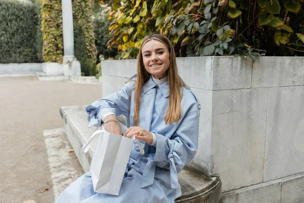 Sorrindo jovem mulher em casaco azul trincheira sentado com saco de compras no banco de concreto no parque verde em Viena — Fotografia de Stock