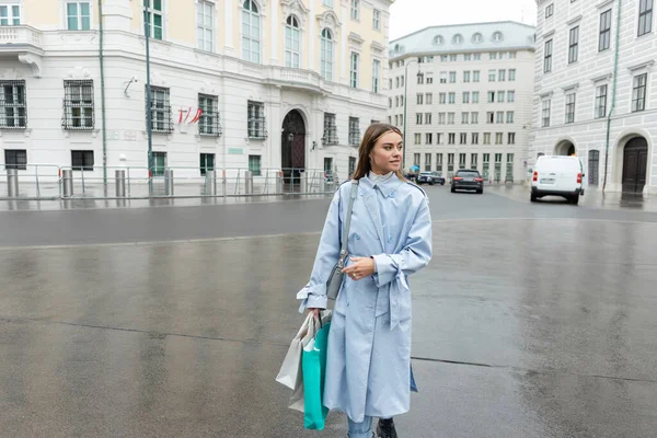 Young woman in blue trench coat walking with shopping bags while walking near buildings in Vienna — Stock Photo