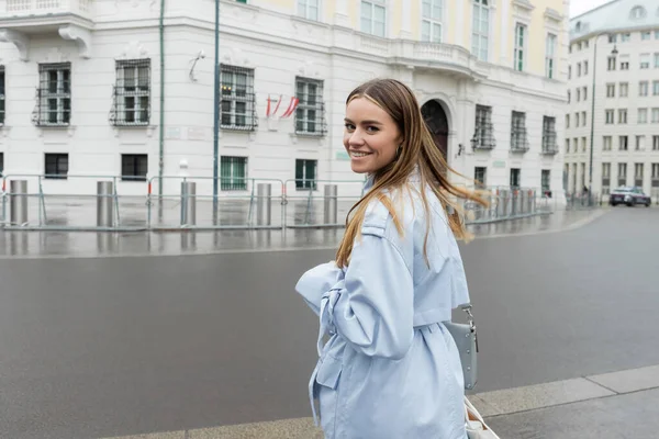 Heureuse jeune femme en trench coat bleu souriant dans la rue urbaine de Vienne — Photo de stock
