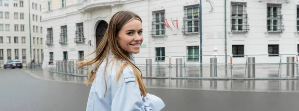 Mujer joven positiva en gabardina azul sonriendo en la calle urbana de Viena, pancarta - foto de stock