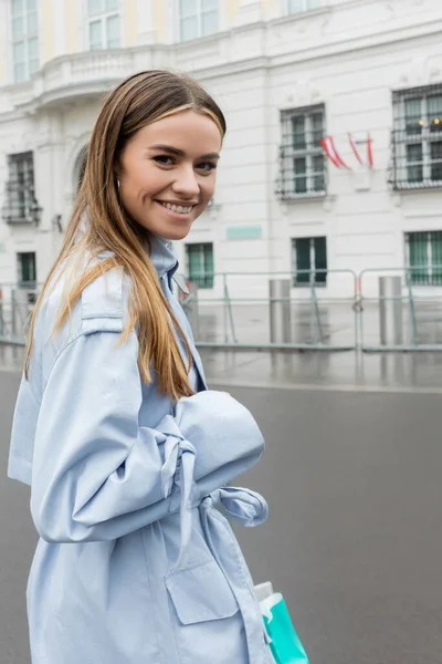 Joyeuse jeune femme en trench coat bleu souriant dans la rue urbaine de Vienne — Photo de stock