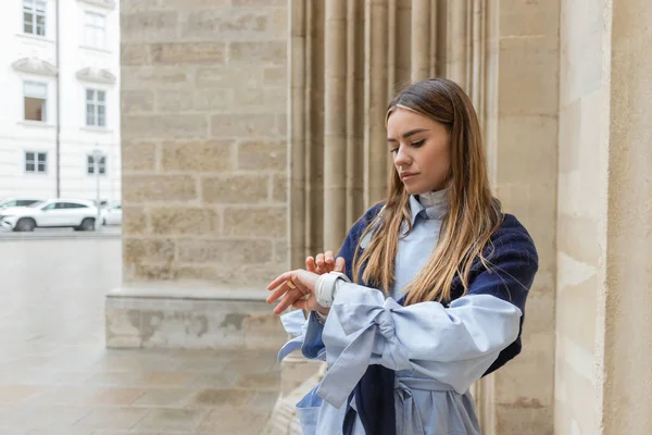 Jeune femme avec écharpe sur le dessus de l'heure de contrôle de trench coat bleu sur montre intelligente près du bâtiment historique à Vienne — Photo de stock